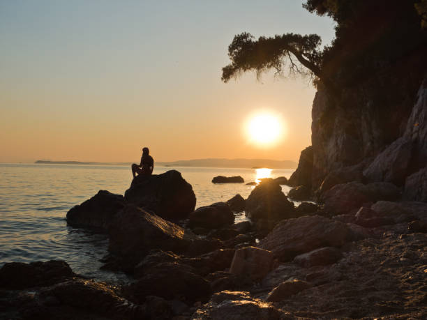 sihouette d’une jeune fille assise sur un rocher au coucher du soleil, la plage de kastani mamma mia, île de skopelos - vibrant color summer rock cliff photos et images de collection