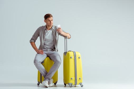 handsome man sitting on yellow suitcase and using smartphone on grey background, travel concept