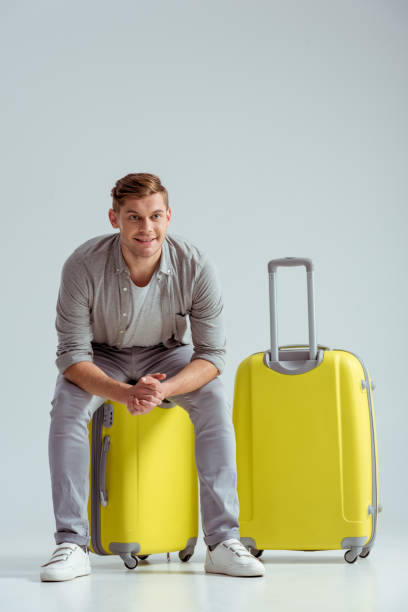 istock smiling handsome man sitting on yellow suitcase with folded hands on grey background, travel concept 1080930896