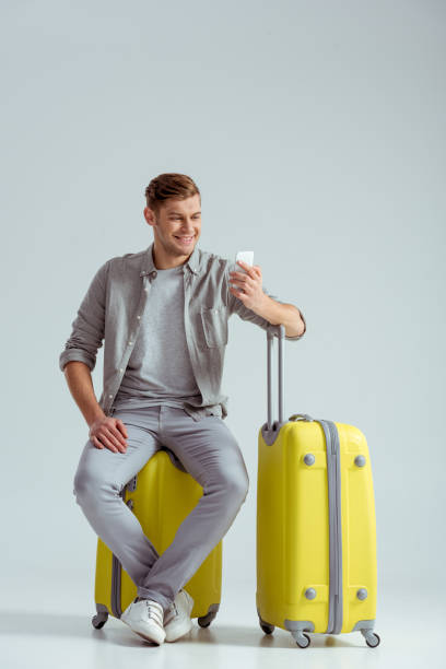 istock smiling man sitting on yellow suitcase and using smartphone on grey background, travel concept 1080930988