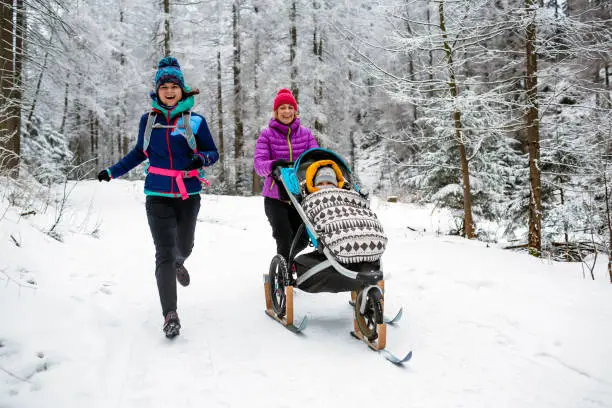 Mother with baby stroller enjoying winter forest with female friend or partner, family time. Hiking and running woman with sledge pram, teamwork concept. Beautiful winter inspirational mountains.