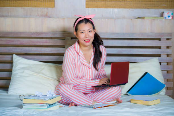 mujer de adolescente joven lindo y feliz nerd asiático chino estudiante en pelo de nerd de la cinta de estudiando en casa habitación sentado en cama con ordenador portátil y libros de texto sonriendo seguro - nerd student female exam fotografías e imágenes de stock