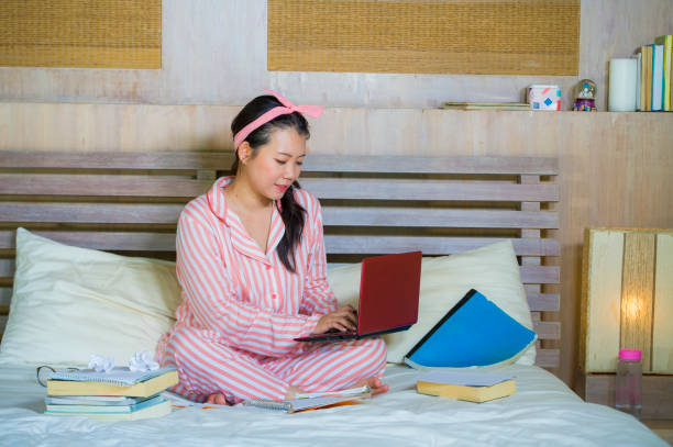 joven linda y feliz asiático americano estudiante adolescente chica nerd en nerd gafas y el pelo de la cinta en hogar habitación sentado en cama con ordenador portátil y libros de texto sonriendo seguro - nerd student female exam fotografías e imágenes de stock