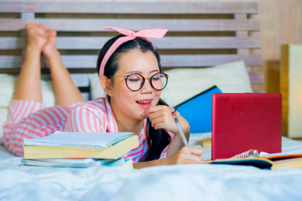 chica adolescente joven lindo y feliz nerd asiático coreano estudiante nerd gafas y pelo de la cinta de estudiando en casa dormitorio acostado en cama con ordenador portátil y libros de texto sonriendo seguro - nerd student female exam fotografías e imágenes de stock