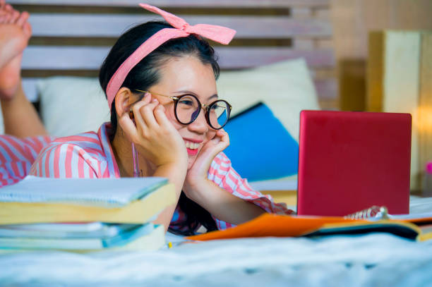 chica adolescente joven lindo y feliz nerd asiático chino estudiante nerd gafas y pelo de la cinta estudiando en casa dormitorio acostado en cama con ordenador portátil y libros de texto sonriendo seguro - nerd student female exam fotografías e imágenes de stock