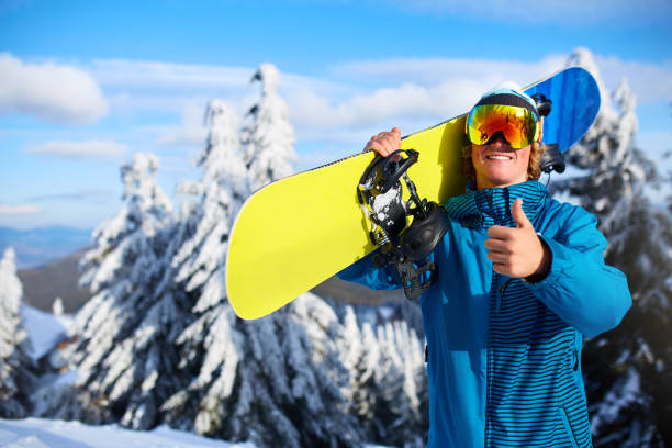 sonriendo el snowboarder posando snowboard llevando en hombros a la estación de esquí cerca de bosque antes de sesión de freeride. jinete mostrando pulgar cartel usa había polarizado gafas. moderno equipo de snowboard. - snowboard non urban scene woods snowboarding fotografías e imágenes de stock