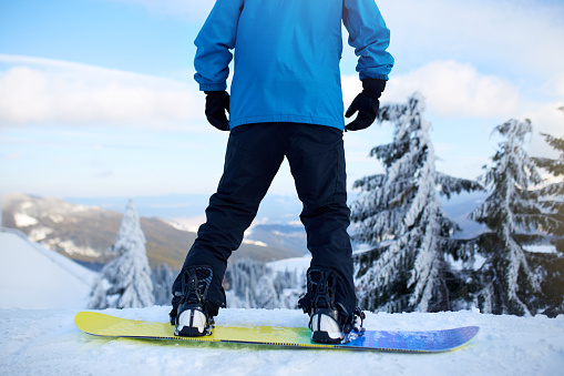 Back view of snowboarder legs on his board before backcountry freeride session in the forest. Man's feet in boots mounted in modern snowboard fast flow bindings fixed with straps. Rider standing at ski resort.