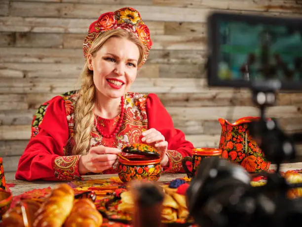 A young woman in traditional Russian clothing, recording a video blog (vlogging) with a camera and Russian breads.