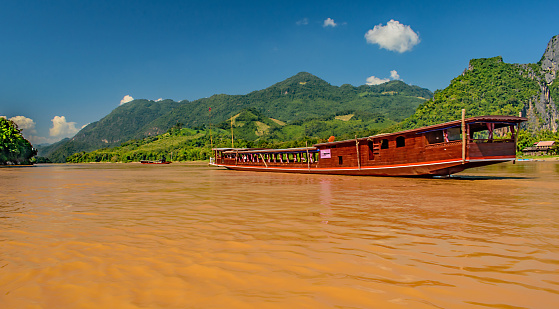 A long brown tour boat sails down the mighty Mekong River in Laos with the lush green mountains lining the shore