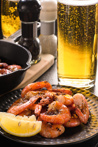 Fried shrimp with lemon on a plate, a glass of beer with bubbles, cast iron frying pan, pepper, salt. Focus on shrimp