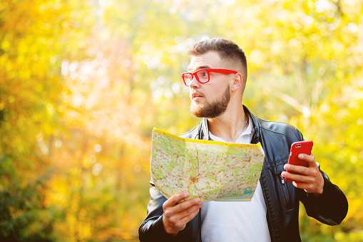 Young white guy with mobile phone and map try find out needed place in a city park. Autumn season