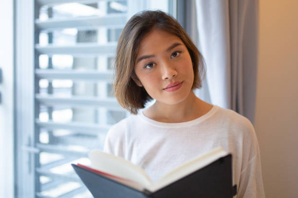 istock Serious young woman reading book at window at home 1081190948