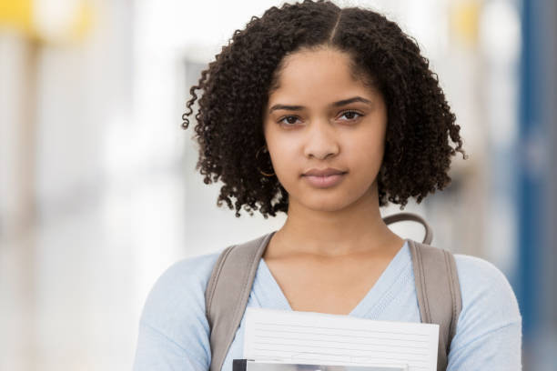 Closeup of female high school student carrying books A serious teenage girl stands in her school wearing a backpack and embracing books and papers.  She stares at the camera with a nervous first day jitters. serious black teen stock pictures, royalty-free photos & images