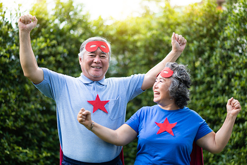 Asian Senior couple in Superhero costume relaxing and celebrating with victory. Elderly people wear red masks and blue shirt with stars having fun, smiling at the park outdoor togetherness.