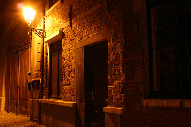 An old lantern shines yellow, seen from below. A strip of blue night sky is visible between the walls of stone houses.