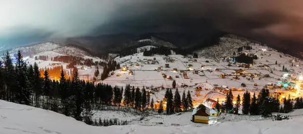 Photo of Snow-capped houses in mountains Carpathians Ukraine. Beautiful sunset over a wide valley. Night Christmas tree in forest. Winter nature. Close up. Landscape. Panorama. Top side view. Slow shutter.