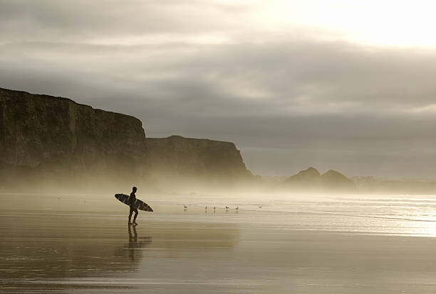winter surfer walking through mist in cornwall winter surfer in walking through mist in cornwall with cliff backdrop near Newquay cornwall england photos stock pictures, royalty-free photos & images