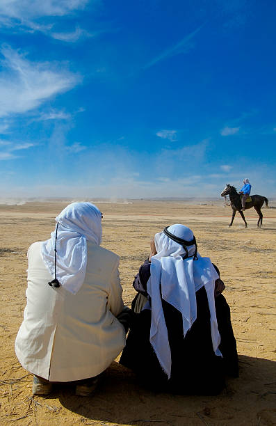 Two teenage boys tourists leap mid-air off a sand dune in the Dubai desert during sunset, dressed in light shirts, shorts, and white socks with headscarves wrapped around their heads. The expansive dune landscape stretches into the distance, bathed in warm golden light as the sun hovers near the horizon. This image captures a moment of pure exhilaration and freedom, blending dynamic motion, desert beauty, and youthful energy during a guided safari experience.The boys feel like they are playing in the giant sandbox.Shot with Canon R5.
