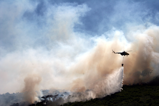 Firefighting helicopter with a bucket suspended on a cable, collecting water from a mountain lake to extinguish the flame in a forest