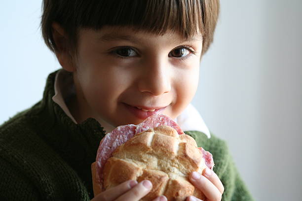 petit garçon enfant souriant manger salami sandwich au pain - elementary age focus on foreground indoors studio shot photos et images de collection