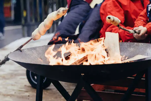 Roasting twist bread on open fireplace. Popular street food and outdoor activity on christmas markets and in winter time in Switzerland. People body parts holding wooden sticks over the fire. People unrecognizable body parts.