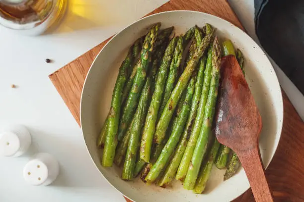 Photo of Top view on roasted asparagus in a white pan on a kitchen table. Modern style, vegetarian food.