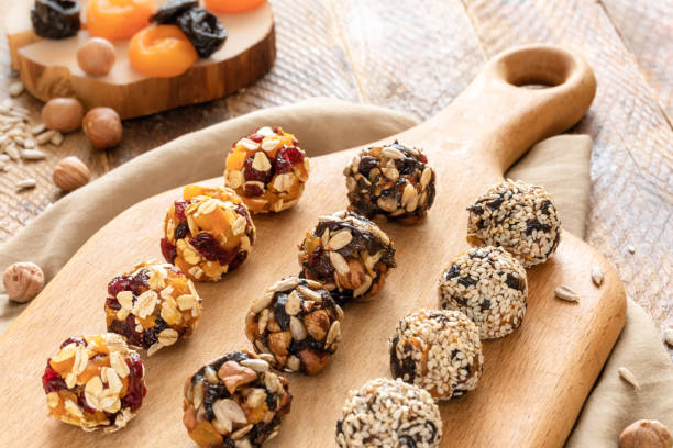 Homemade energy balls are lying in rows on kitchen cutting board on wooden table. Homemade different kinds of energy balls made from pieces of dried cranberries, apricots, prunes, sunflower seeds, hazelnuts, oat flakes are lying in rows on kitchen cutting board and ingredients for their preparation on wooden table. Healthy organic sweets. plasma ball stock pictures, royalty-free photos & images