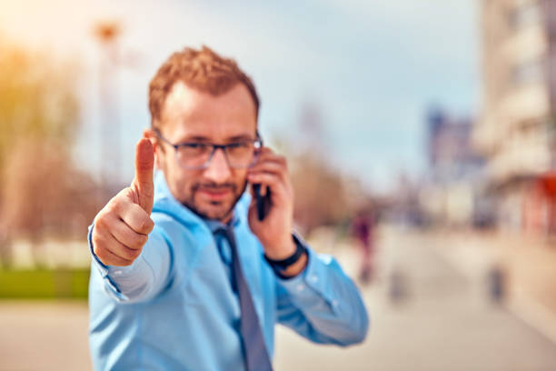 Funny businessman with thumbs up while using smartphone outdoors.