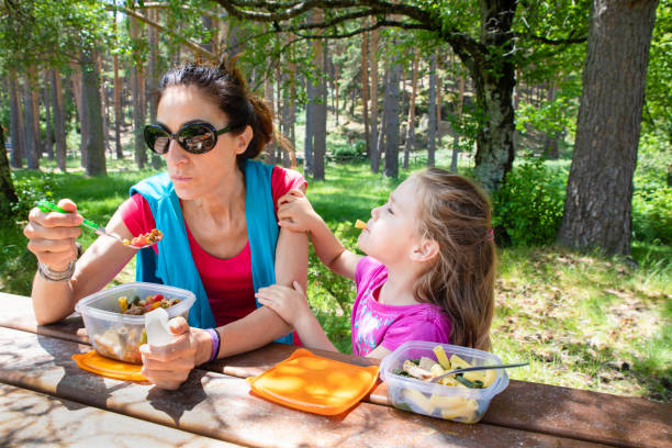 funny girl and woman eating pasta salad from a plastic lunchbox in a table picnic in the country funny scene: four years old blonde girl making fun playing with macaroni, looking at her mother, eating pasta from a plastic lunchbox in a table picnic in the country kids eating tuna stock pictures, royalty-free photos & images