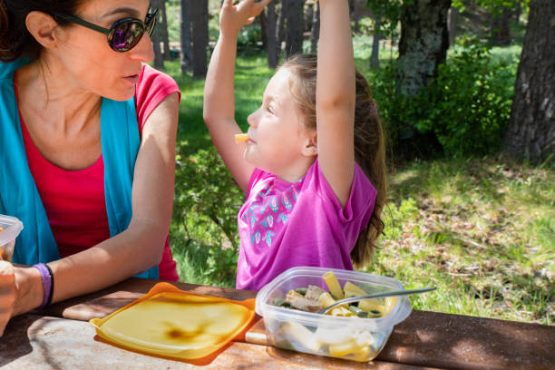 funny girl playing with macaroni in her mouth looking at her mother eating from a plastic lunchbox in a table picnic in country funny four years old blonde girl making fun playing with macaroni, looking at her mother, eating pasta from a plastic lunchbox in a table picnic in the country kids eating tuna stock pictures, royalty-free photos & images