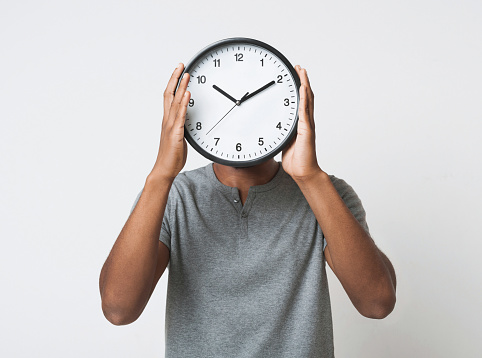 Time management concept. Black man covering his face with big clock on white background