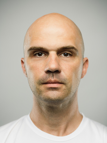Close up portrait of mid adult adult caucasian man with blank expression against gray background. Vertical shot of real serbian man staring in studio with bald shaved head and brown eyes. Photography from a DSLR camera. Sharp focus on eyes.