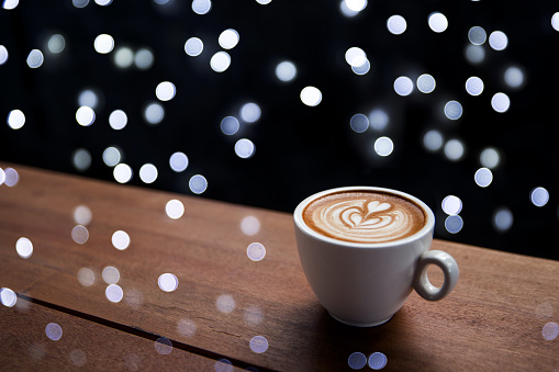 Tasty cappuccino with some blurred lights from Christmas tree on wooden background. Place for text. Holiday concept.