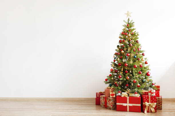 istock Big beautiful christmas tree decorated with beautiful shiny baubles and many different presents on wooden floor. White wall background with a lot of copy space for text. Close up. 1081416706