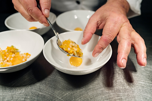 Chef carefully spooning in a selection of delicately cooked vegetables, leeks, carrots and celery along with a smoked egg yolk into a bowl before the consommé is added. Colour, horizontal with some copy space. Photographed on location in a restaurant on the island of Møn in Denmark.