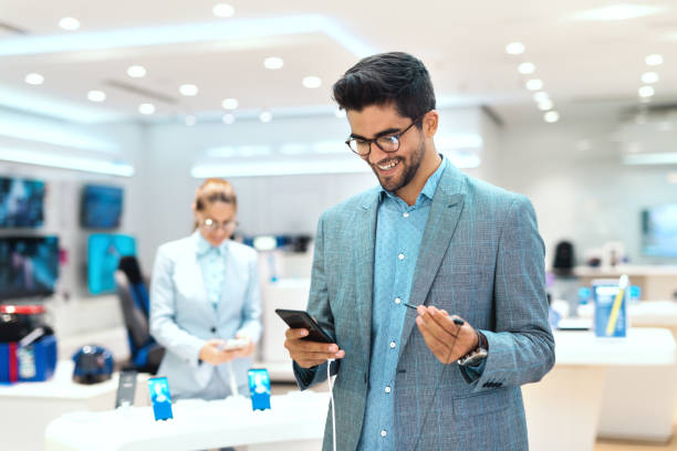 istock Young mixed race man dressed smart casual and with eyeglasses trying out smart phone. Tech store interior. 1081447480