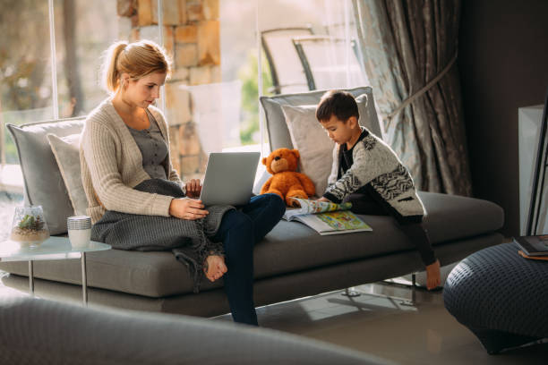 Mother working on laptop and son reading story book Woman working on laptop with her son reading a storybook. Mother looking busy working on laptop and little boy with a picture book at home. working at home with children stock pictures, royalty-free photos & images