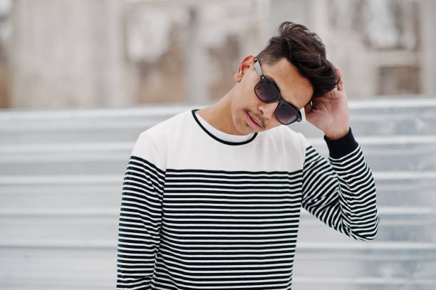 Fashionable handsome model man in a T-shirt and white pants with black shoes near the glossy metal wall