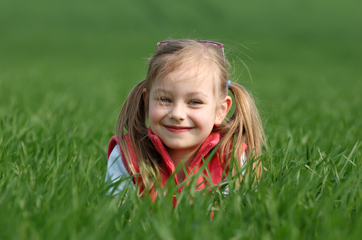 Portrait of a beautiful brunette girl looking at the camera and smiling, standing on the street against the backdrop of greenery on a summer day. Young curious child raises his eyes and lowered his head looks at the camera close-up outdoors. Face Eyes Contemplative Child.