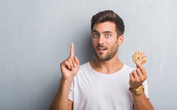 istock Handsome young man over grey grunge wall eating chocolate chip cooky surprised with an idea or question pointing finger with happy face, number one 1081504580