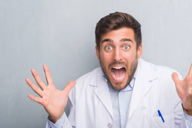 istock Handsome young professional man over grey grunge wall wearing white coat celebrating crazy and amazed for success with arms raised and open eyes screaming excited. Winner concept 1081505908