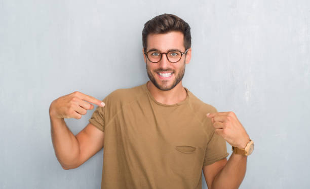 istock Handsome young man over grey grunge wall wearing glasses looking confident with smile on face, pointing oneself with fingers proud and happy. 1081507030