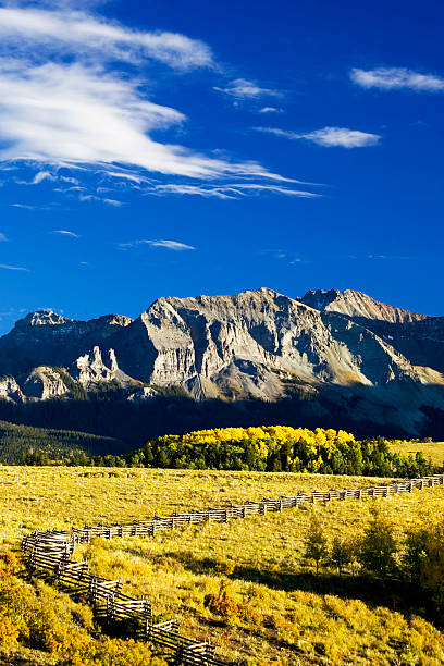 A wooden fence sits on a grassy hillside overlooking a mountain range. The sky is clear and blue, and the mountains are covered in snow. The scene is peaceful and serene