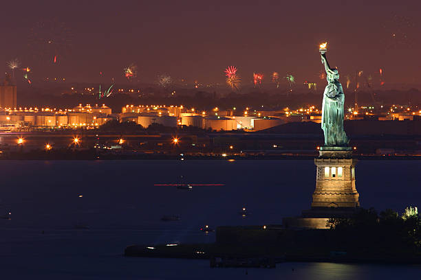 A photograph of the Statue of Liberty illuminated by fireworks.  The Statue of Liberty is seen from a worm's eye point of view.  The only visible parts of the statue are the shoulders, face and arm, and the statue enters the frame from the bottom right corner.  The arm extends to the exact mid-point of the frame, with the torch illuminated by the surrounding fireworks.  The fireworks burst in shades of yellow, orange and red, and they fill the sky with their bright, graphic patterns.  The sky itself is pitch black.