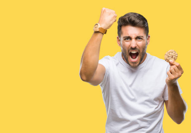 istock Young handsome man eating chocolate chips cookie over isolated background annoyed and frustrated shouting with anger, crazy and yelling with raised hand, anger concept 1081515122