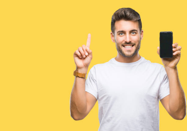 istock Young handsome man showing smartphone screen over isolated background surprised with an idea or question pointing finger with happy face, number one 1081515520