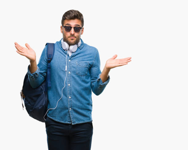 young handsome tourist man wearing headphones and backpack over isolated background clueless and confused expression with arms and hands raised. doubt concept. - head and shoulders audio imagens e fotografias de stock