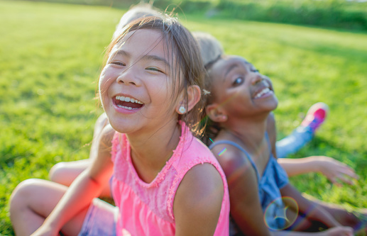 Three elementary school girls sit on the grass in a park, laughing.