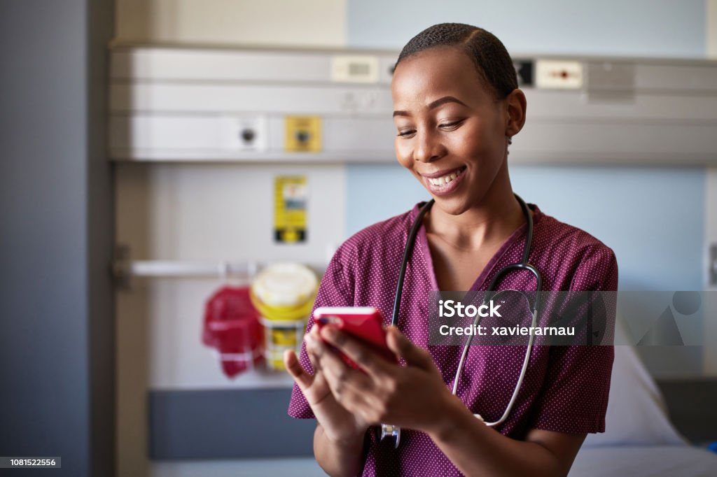 Smiling nurse with smart phone at hospital ward Smiling nurse text messaging on smart phone. Young medical professional is wearing uniform. She is in hospital ward. Mobile Phone Stock Photo