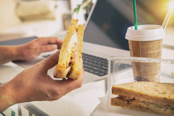 Woman eating a breakfast sandwich and drinking coffee while working with a laptop